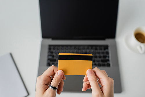 top view of woman holding credit card near blurred laptop with blank screen, notebook and cup of coffee with saucer on white table, modern workspace, work from home, digital payment