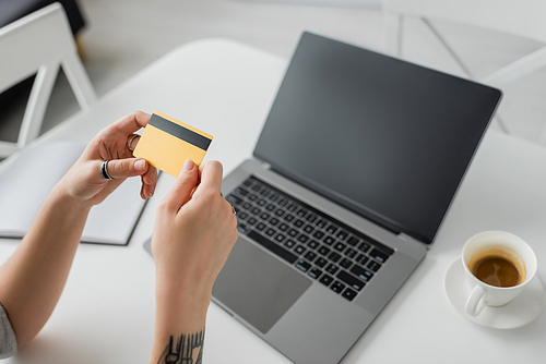 high angle view of tattooed woman holding credit card near blurred laptop with blank screen, notebook and cup of coffee with saucer on white table, modern workspace, work from home, digital payment