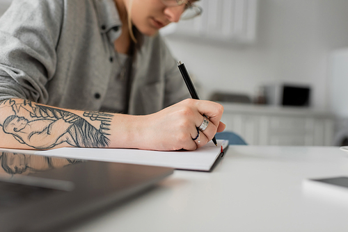 cropped view of young woman with tattoo on hand writing in notebook, taking notes, having inspiration while holding pen near laptop on white table, blurred foreground, work from home