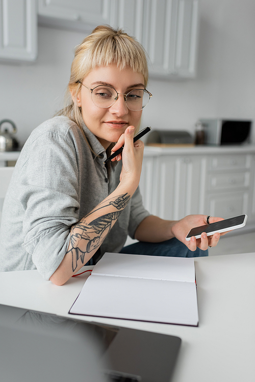 happy young woman with tattoo on hand and bangs holding smartphone with blank screen and pen near notebook and laptop on white table, blurred foreground, work from home