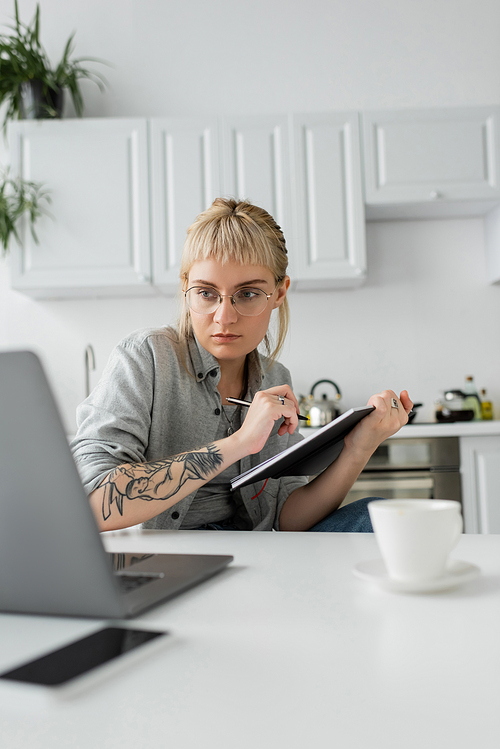 young woman with tattoo on hand and bangs holding notebook, taking notes near smartphone and laptop on white table, blurred foreground, work from home