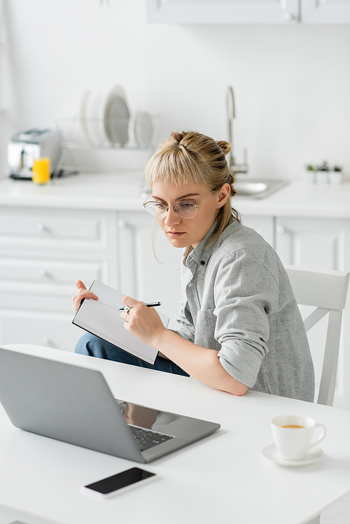 young woman with tattoo on hand and bangs holding notebook, taking notes near smartphone and laptop on white table, blurred background , work from home