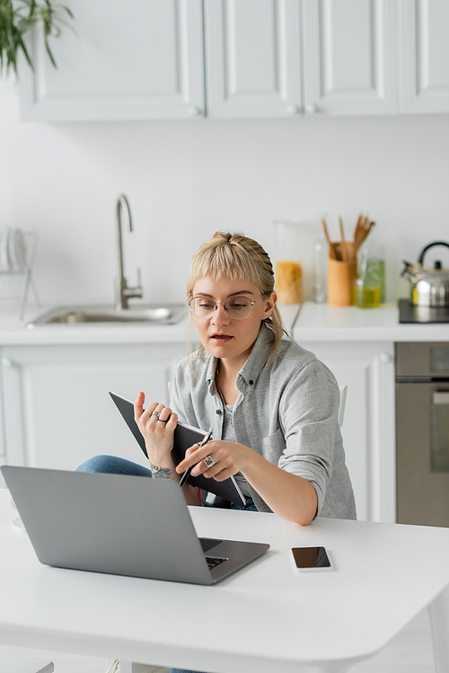 young woman with tattoo on hand and bangs holding notebook, near smartphone with blank screen and looking at laptop on white table, blurred foreground, work from home