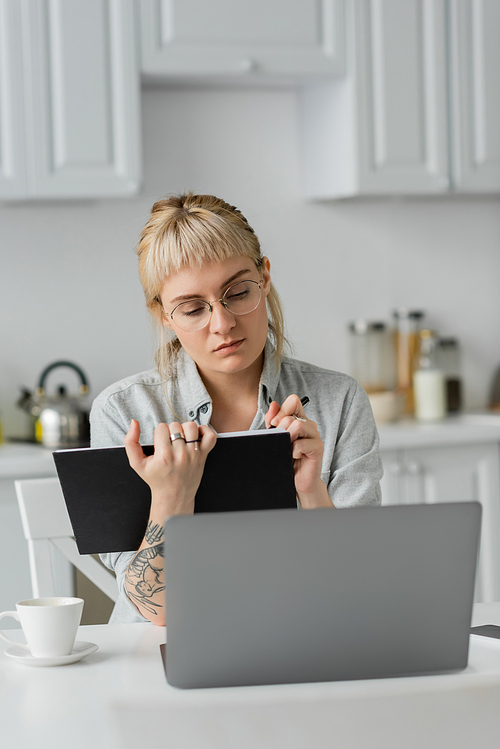 young woman in eyeglasses with tattoo on hand and bangs holding notebook, taking notes, sitting near laptop and cup of coffee on white table, blurred background, work from home