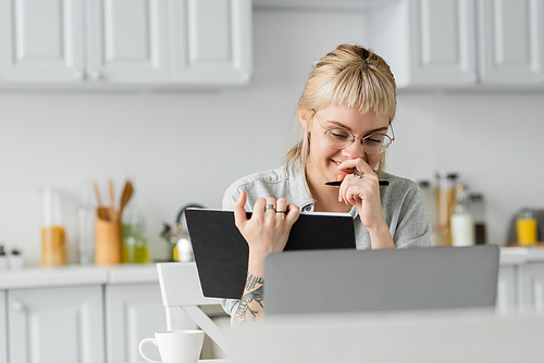 happy young woman in eyeglasses with tattoo on hand and bangs laughing, covering mouth, holding notebook, taking notes, sitting near laptop and cup of coffee on table, blurred background,