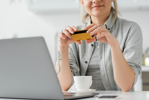 cropped view of happy young woman with tattoo on hand holding credit card, sitting near laptop, smartphone and cup of coffee on white table, blurred background, work from home