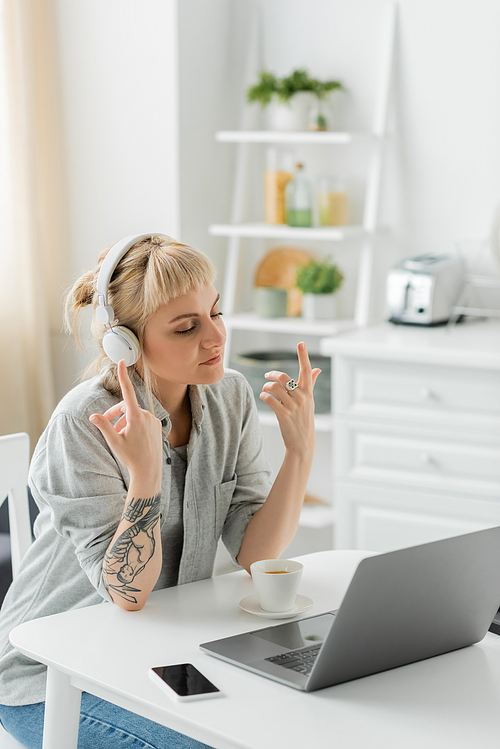 young woman with bangs and tattoo on hand sitting in wireless headphones and gesturing near laptop, cup of coffee and blurred smartphone with blank screen on table, freelance, work from home