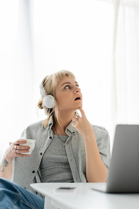 pensive young woman with blonde hair, bangs and tattoo on hand sitting in wireless headphones and holding cup of coffee near laptop and blurred smartphone on table. freelance. work from home