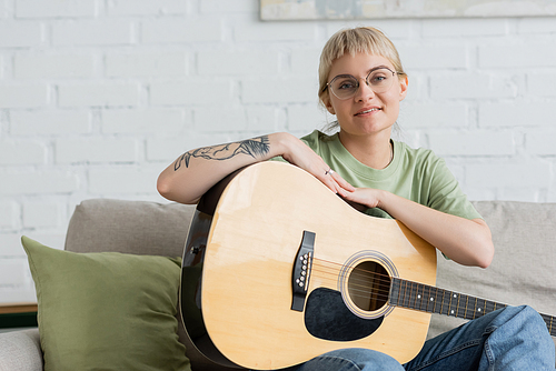 pleased young woman in glasses with bangs and tattoo on hand holding acoustic guitar and looking at camera while sitting on comfortable couch in modern living room at home