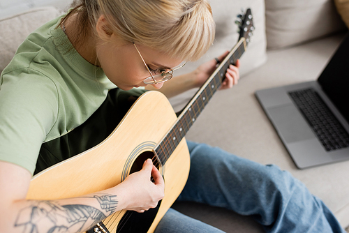 young woman in glasses with bangs and tattoo holding acoustic guitar and learning how to play near laptop and sitting on comfortable couch in modern living room at home, virtual lessons