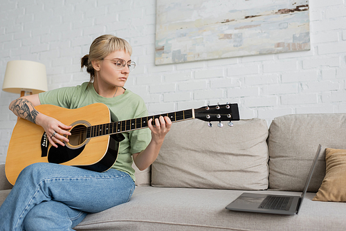 young woman in glasses with bangs and tattoo playing acoustic guitar while looking video tutorial on laptop and sitting on comfortable couch in modern living room at home, skill development