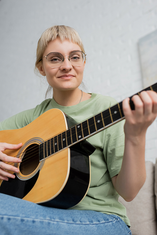 happy young woman in glasses with bangs playing acoustic guitar and sitting on comfortable couch in modern living room, learning music, skill development, music enthusiast