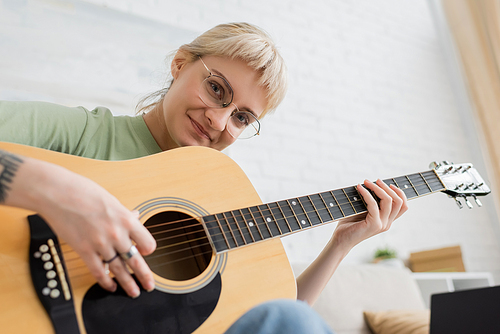 cheerful young woman in glasses with bangs and tattoo on hand playing acoustic guitar and looking at camera while sitting in modern living room, learning music, music enthusiast
