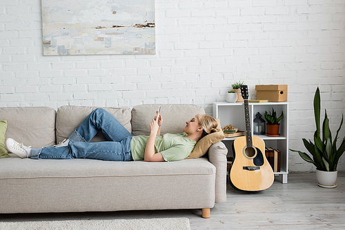 side view of happy young woman with blonde and short hair, bangs and eyeglasses using smartphone while resting on comfortable couch near guitar in modern living room with painting on wall and plants