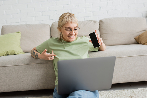 cheerful and tattooed woman with bangs and eyeglasses using laptop while sitting on carpet and holding smartphone with blank screen near comfortable couch in modern living room