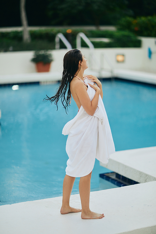 young brunette woman with wet hair wrapped in white towel standing next to outdoor swimming pool with shimmering water in Miami, summer getaway, youth, poolside relaxation, vacation mode