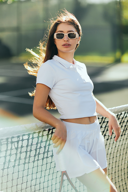 Miami, Florida, active lifestyle, beautiful young woman standing in stylish outfit and sunglasses while holding racket near tennis net, blurred background, iconic city, sunny day, vacation