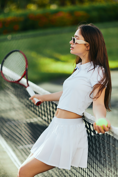 tennis court in Miami, athletic young woman with long hair standing in white outfit and sunglasses while holding blurred racket and ball and leaning on tennis net, green background, iconic city