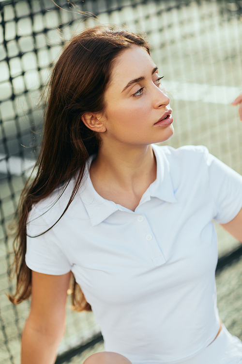 portrait of attractive young woman with long brunette hair wearing white polo shirt and looking away after training on tennis court, tennis net on blurred background, Miami, Florida