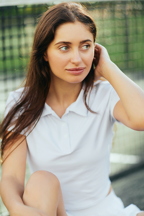 portrait of attractive young woman wearing white polo shirt and looking away while adjusting long brunette hair after training on tennis court, tennis net on blurred background, Miami, Florida
