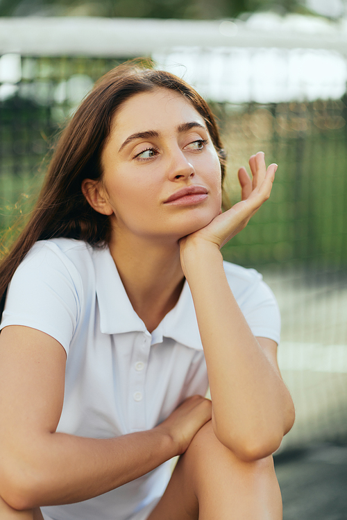 tennis court in Miami, portrait of dreamy female tennis player with brunette hair wearing white polo shirt and looking away after training, tennis net on blurred background, Florida