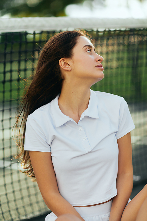 tennis court in Miami, portrait of distracted female tennis player with brunette hair wearing white polo shirt and looking away after training, tennis net on blurred background, Florida
