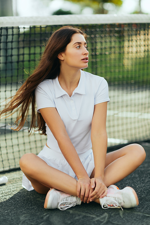 distracted female player on tennis court, young woman with long hair sitting with crossed legs in white outfit and sneakers and looking away near tennis net, blurred background, Miami, downtime
