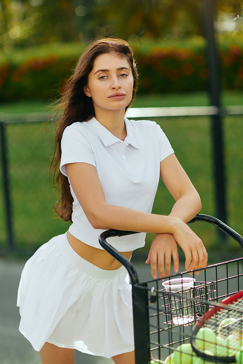 healthy habits, sporty young woman with brunette hair standing in white outfit with skirt and polo shirt and leaning on cart with balls and racket, blurred background, tennis court in Miami