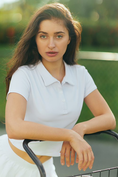 pretty tennis player, sporty young woman with brunette hair standing in white polo shirt near tennis cart, blurred green background, looking at camera, tennis court in Miami
