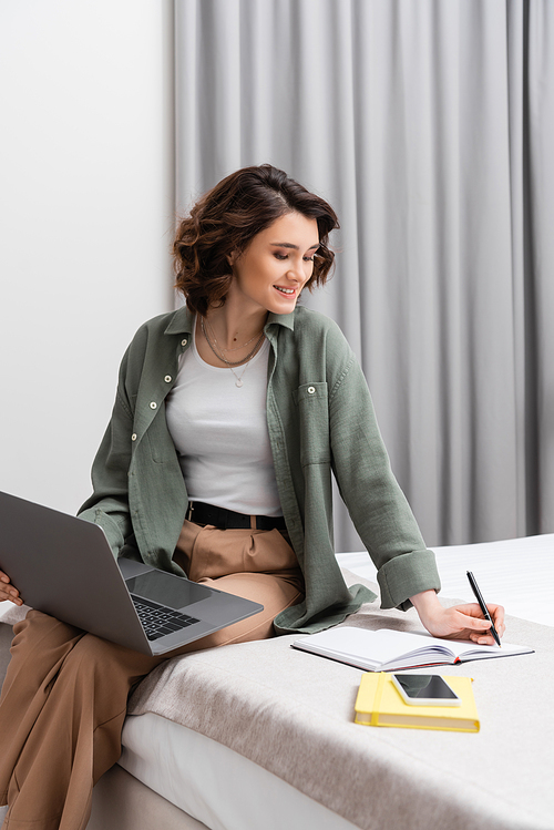 pleased young woman with wavy brunette hair sitting on bed with laptop and writing in notebook near smartphone with blank screen, notepad and grey curtains in comfortable hotel room