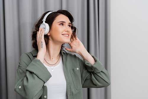 delighted young woman in casual clothes, with tattoo and with wavy brunette hair looking away while listening music in wireless headphones near grey curtains in modern hotel room, weekend getaway