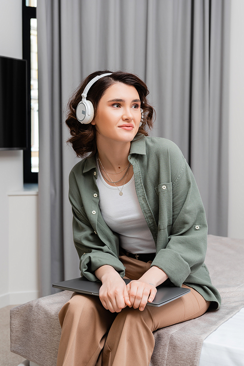 pleased woman with wavy brunette hair, in casual clothes and wireless headphones listening audio lesson while sitting on bed with laptop near grey curtains in modern hotel room, study and travel