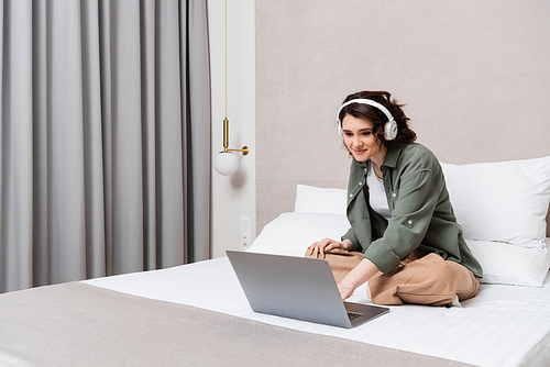 happy young woman in casual clothes and wireless headphones sitting on bed near white pillows, grey curtains and wall sconce while watching movie on laptop in hotel room, leisure and travel