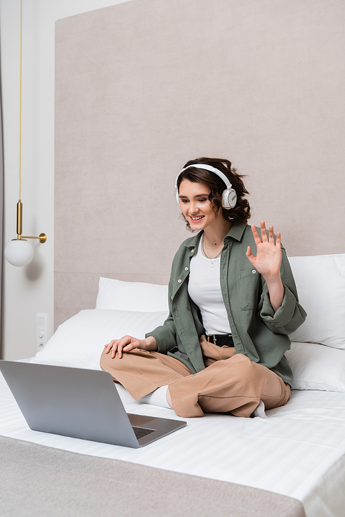 overjoyed woman with wavy brunette hair, in wireless headphones and casual clothes sitting on bed and waving hand during video call on laptop near white pillows and wall sconce in hotel room