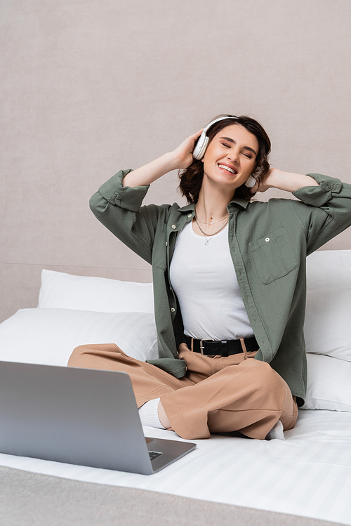 carefree woman with wavy brunette hair and closed eyes listening musical podcast in wireless headphones while sitting on bed near laptop and white pillows in hotel suite, leisure and travel