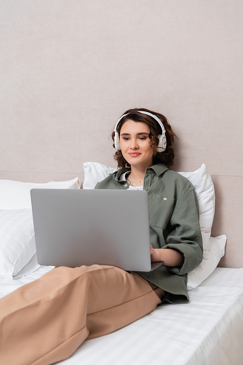 young woman with wavy brunette hair, in casual clothes and wireless headphones sitting on bed near white pillows and smiling while watching film on laptop in hotel room