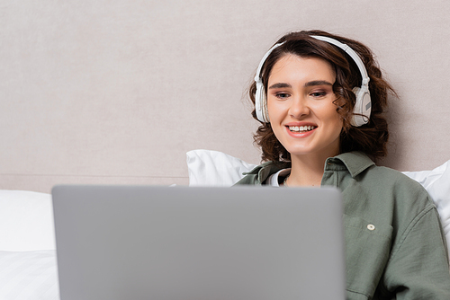 young and cheerful woman with wavy brunette hair watching film on blurred laptop while sitting in wireless headphones near grey wall and white pillows in modern hotel room