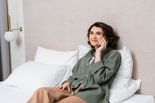 pleased young woman in casual clothes, with wavy brunette hair talking on mobile phone near grey wall and white pillows on comfortable bed in model hotel room, leisure and travel