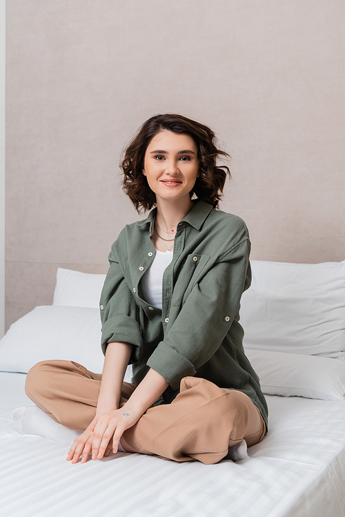 carefree woman with wavy brunette hair sitting on bed in casual clothes, with crossed legs, looking at camera near white pillows and grey wall in contemporary hotel suite, leisure and travel