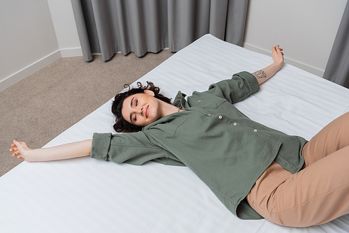 high angle view of young and carefree woman with wavy brunette hair, tattoo and closed eyes smiling while lying on bed in casual clothes in modern and cozy hotel suite, leisure and travel