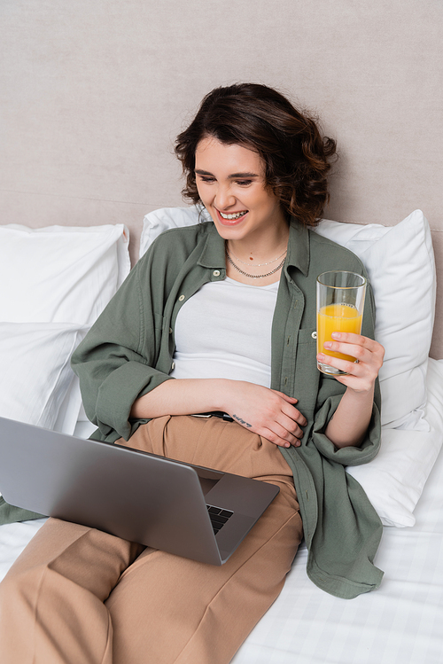 young and carefree woman in casual clothes, with wavy brunette hair and tattoo holding glass of fresh orange juice while watching movie on laptop on bed near white pillows and grey wall in hotel room