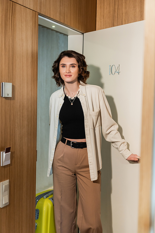 delighted woman in stylish clothes, white shirt and beige pants, with wavy brunette hair, holding yellow suitcase and opening door of modern hotel room with secure entry and keyboard access