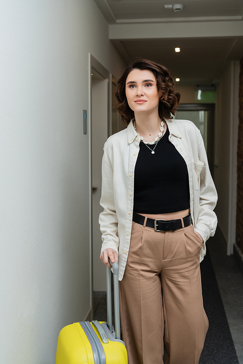 young woman with wavy brunette hair, in black crop top, white shirt and beige pants walking with hand in pocket and yellow suitcase along corridor in contemporary hotel