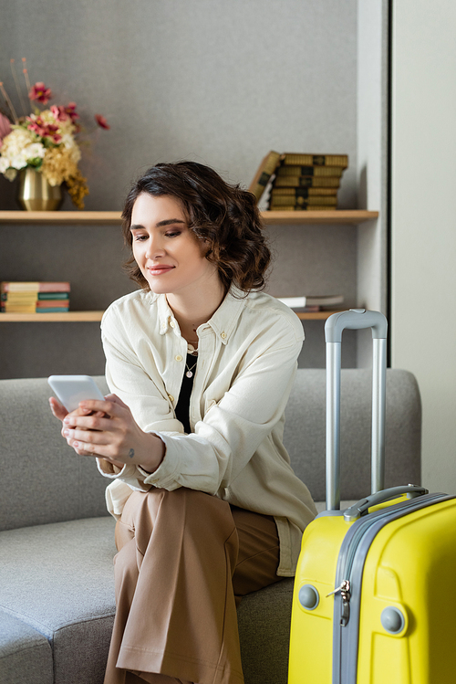 delighted tattooed woman with wavy brunette hair sitting on couch near yellow travel bag and browsing internet on smartphone near books and vase with flowers on blurred background in hotel lobby