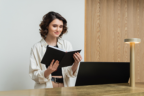 positive receptionist in trendy casual clothes, with wavy brunette hair standing with notebook near computer monitor and lamp on front desk in lobby of contemporary hotel