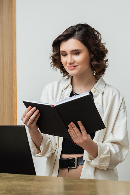 cheerful concierge in trendy casual clothes and with wavy brunette hair looking at notebook near computer monitor while working at reception desk in contemporary hotel, digital nomad