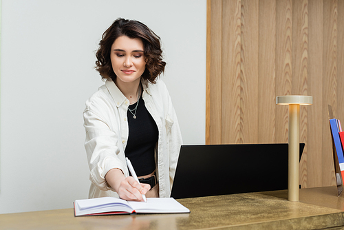 friendly concierge in stylish casual clothes, with wavy brunette hair writing in notebook near computer monitor and lamp while working at reception desk in lobby of contemporary hotel