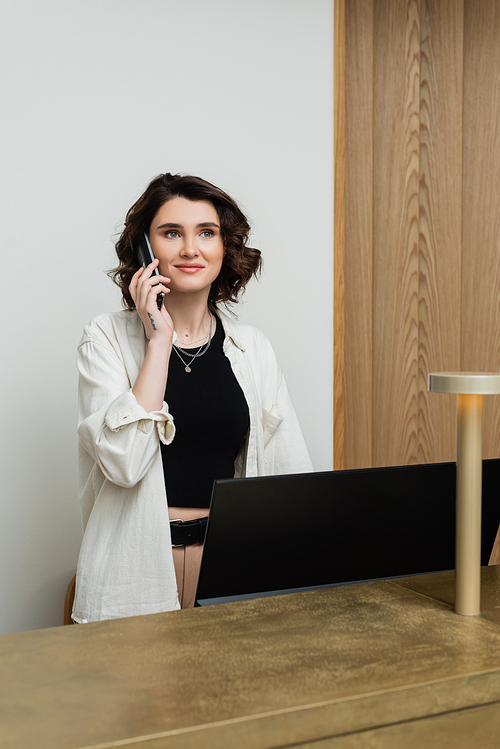 happy concierge with wavy brunette hair and tattoo wearing stylish clothes and talking on telephone at reception desk near computer monitor and lamp in lobby of modern hotel, customer service