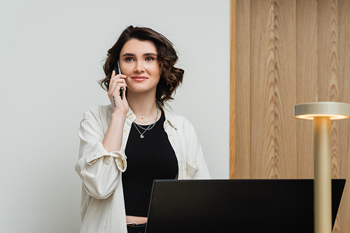 pleased young woman in stylish casual clothes, with wavy brunette hair and tattoo talking on telephone while working as receptionist at front desk near computer monitor and lamp in hotel lobby