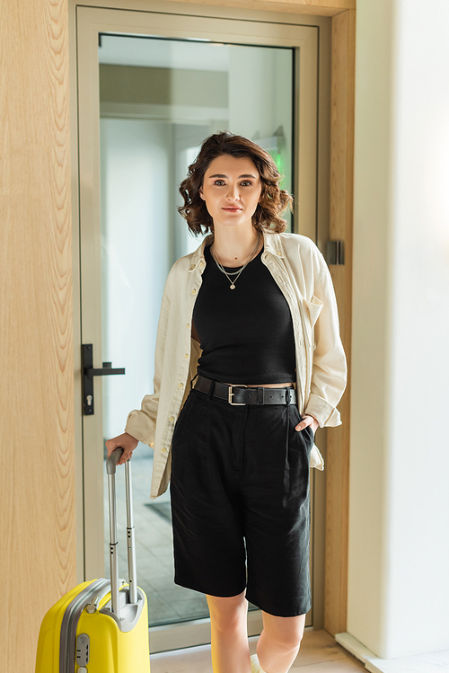 fashionable and self-assured woman in white shirt, crop top and black shorts standing with hand in pocket near yellow suitcase and glass door while looking at camera in lobby of modern hotel